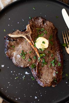 two steaks with butter and parsley on a black plate next to a fork