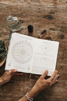 two hands holding an open book on top of a wooden table next to other items