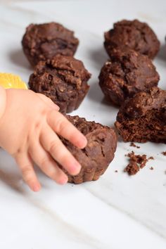 a baby reaching for some chocolate cookies on a white surface with other muffins in the background