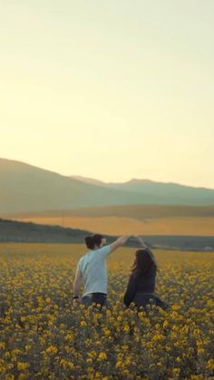 two people standing in a field with yellow flowers and mountains in the background at sunset