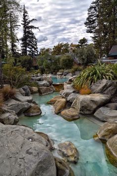 a river running through a lush green forest next to rocks and trees on a cloudy day