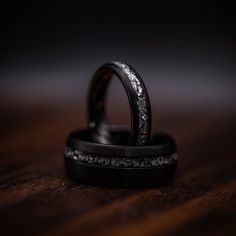 two black wedding rings sitting on top of each other in front of a dark background