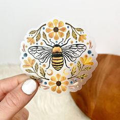 a hand holding up a sticker with a bee on it's side and flowers in the middle