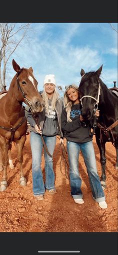 three women standing next to two horses on a dirt field with trees in the background