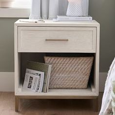 a white night stand with a basket and lamp on it next to a window in a bedroom