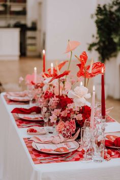 a long table is set with red and white place settings, candles and flowers in vases