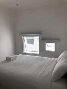 a bed with white sheets and pillows in a room next to two windows that have blind shades on them