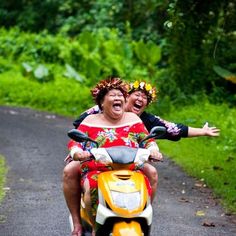 two people riding on the back of a yellow scooter in front of trees