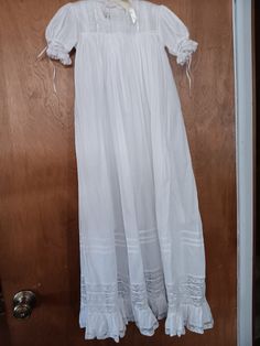 This is a Baptismal or a Christening gown.  It is made with white cotton baptist, the yoke is made up of lace, beading lace, and white ribbon.  The collar is made of lace.  The gown has short sleeves with a lace and ribbon band at the bottom of the sleeves.  At the bottom of the gown is a 3.5 inch lace band and a 3 inch ruffle and lace band. Above the lace band are three rows of pleats.  The gown is 32 inches from the yoke to the bottom of the gown.  The gown had entradau where the sleeves are attached to the yoke, where the beading lace is attached to the sleeve, where the lace is attached to the sleeve beading lace, where the skirt is attached to the skirt, where the lace is attached to the neck, where the bottom lace band is attached to the skirt and where the ruffle band is attached to White Long Sleeve Baptism Dress With Lace Trim, Classic Lace Baptism Dress With Lace Trim, Classic Baptism Dress With Lace Trim, Vintage Lace Trim Baptism Dress, Vintage Cotton Baptism Dress With Lace Trim, Lace Bands, Christening Gowns, French Seam, White Ribbon