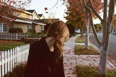 a woman standing on the sidewalk in front of a white picket fence and trees with red leaves