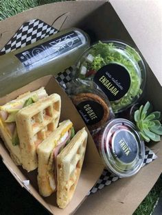 a box filled with sandwiches and salads on top of a grass covered field next to a bottle of booze