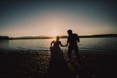 a man and woman holding hands on the shore of a body of water at sunset