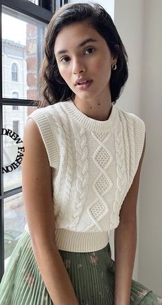 a woman is sitting by a window wearing a white sweater and green pleated skirt