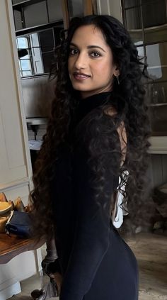 a woman with long curly hair standing next to a counter in a kitchen, looking at the camera