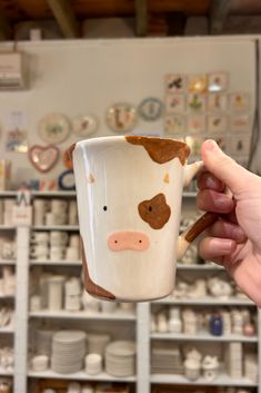 a hand holding a coffee mug with a cow painted on it in front of shelves