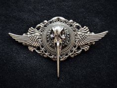 A captivating hand-crafted metal alloy badge brooch in silver tones featuring a central miniature bird skull.  An elegant gift for any stylish Goth or Steampunk gentleman or lady to wear and enjoy. A silver-winged filigree silver base is topped with silver cogs and a silver miniature bird skull. The filigree base has a silver brooch pin measuring approximately 2.5cms/1" wide on the reverse side (see example in picture). This striking eye-catching item has been individually designed and handmade Cogs And Gears, Steampunk Bird, Steampunk Goth, Raven Bird, Skull Pin, Steampunk Hat, Silver Brooch Pin, Small Bees, Raven Skull