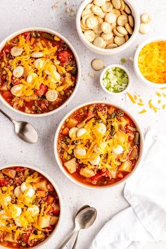 three bowls filled with chili and cheese next to two spoons on a white surface