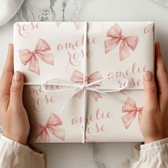 two hands holding a wrapped gift box with pink bows on it and white wrapping paper