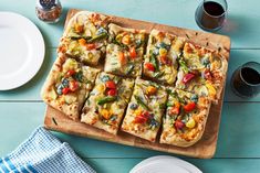 a wooden cutting board topped with sliced pizza on top of a blue table next to plates and cups