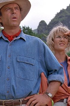 a man and woman standing next to each other in front of some mountains with trees