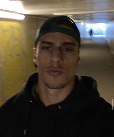 a man standing in a parking garage next to a yellow wall and wearing a green hat