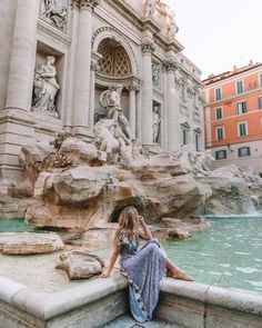 a woman sitting on the edge of a fountain in front of a building with statues