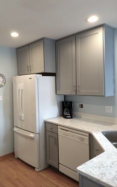 a kitchen with gray cabinets and white counter tops is pictured in this image, there are two clocks on the wall above the refrigerator