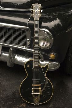 an electric guitar sitting on the ground next to a black and gold car with it's hood up