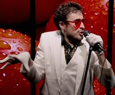 a man in a white suit and red glasses singing into a microphone with his hands out