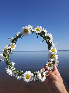 a hand holding up a wreath made out of daisies on the beach with water in the background