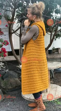 a woman wearing a yellow crocheted vest and brown boots standing in front of a tree