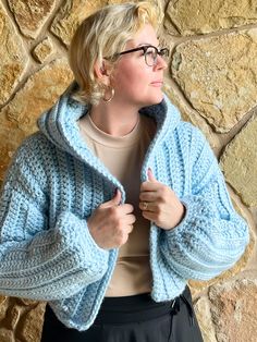 a woman standing in front of a stone wall wearing a blue sweater and black pants