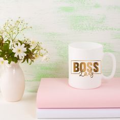 a white coffee mug sitting on top of a pink book next to a vase with flowers