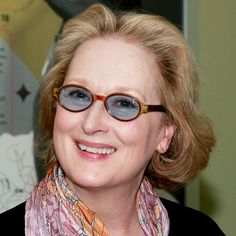 a woman wearing glasses and a scarf smiles at the camera while standing in front of a painting