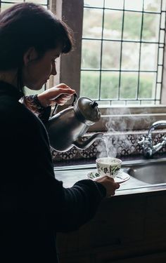 a woman pouring tea into a cup in front of a kitchen sink with the window open