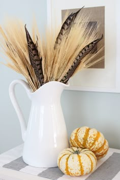 two pumpkins sitting on a table next to a white pitcher filled with dry grass