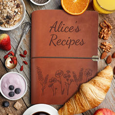 an open leather recipe book surrounded by fruits, nuts, and other foods on a wooden table