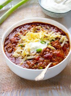 a white bowl filled with chili and cheese next to bowls of sour cream, green onions and celery