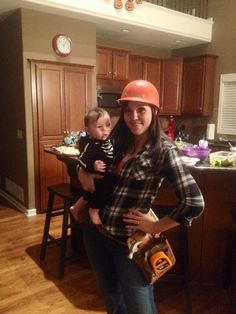 a woman holding a baby wearing a hard hat while standing in front of a kitchen counter