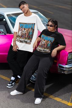 two people sitting on the hood of a car in front of a pink and black car