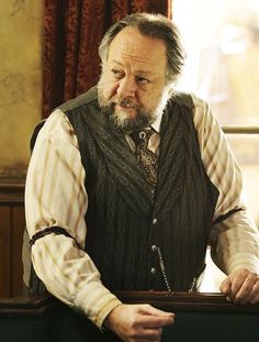 an older man wearing a vest and holding a knife in his hand while standing next to a window