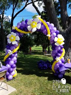 a purple and yellow balloon arch with flowers on it in the middle of a lawn