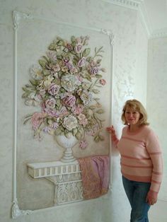 a woman standing in front of a wall with flowers on it and a painting behind her