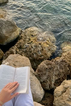 a person sitting on rocks reading a book by the water's edge with their hand resting on an open book