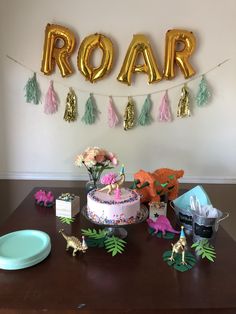 a birthday cake and decorations on a table in front of a roar sign with balloons