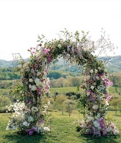 an arch covered in flowers on top of a lush green field with trees and hills in the background