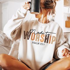 a woman sitting on a couch holding a coffee mug in her hand and wearing a sweatshirt that says made to worship