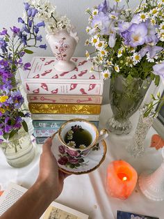 a person is holding a tea cup in front of some flowers and books on a table