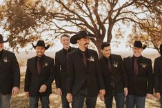 a group of men standing next to each other in front of a tree wearing black suits and hats