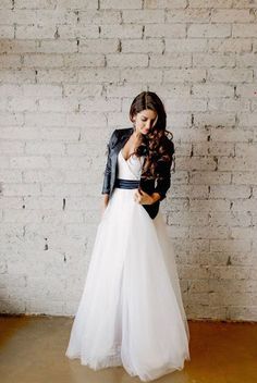 a woman standing in front of a brick wall wearing a white dress and black jacket